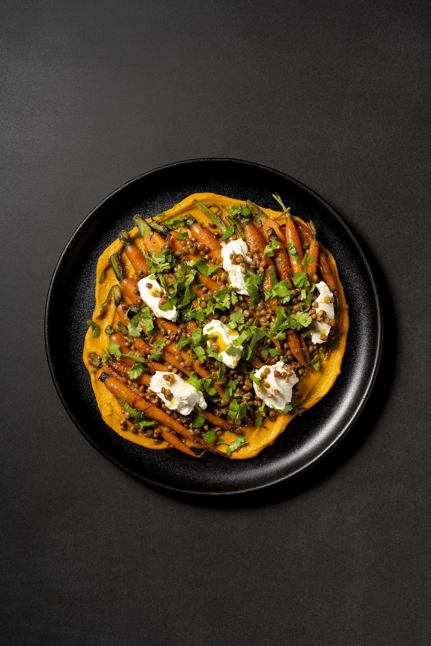 A dark plate with pumpkin puree topped with carrots herbs, nuts, and dollops of cream.